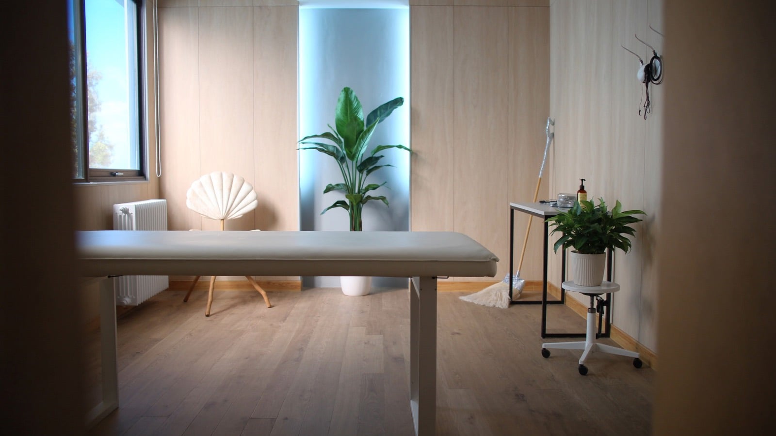 The Pearl Coast physiotherapy treatment room: a low timber treatment table and a shell-shaped chair beside a tall green plant, with timber floor and walls lit by soft daylight from a window.